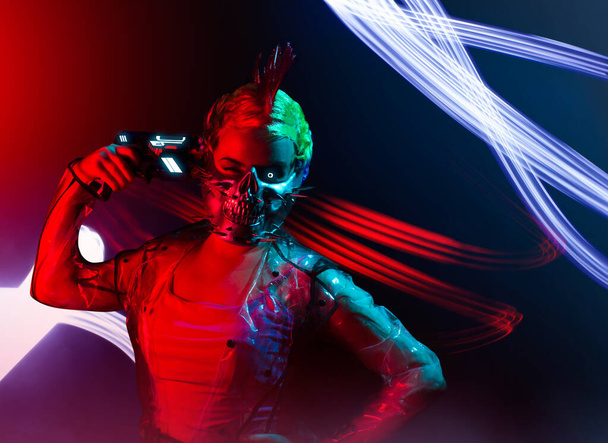 Cyborg punk woman with mohawk hairstyle in spiked mask and transparent raincoat putting a gun to her head. - Foto, afbeelding