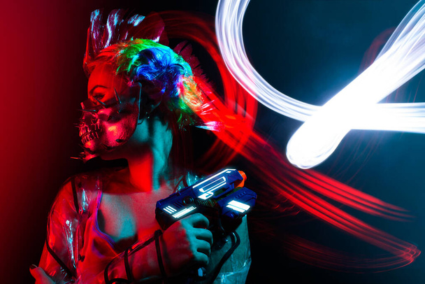 Όμορφη γυναίκα cyberpunk με μοχόκ χτένισμα σε αιχμηρή μάσκα κρατώντας ένα πιστόλι. - Φωτογραφία, εικόνα