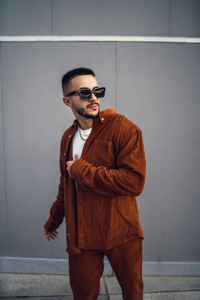 A vertical shot of a young handsome European man in a brown stylish outfit and sunglasses posing at the camera - Foto, Bild