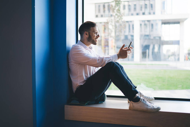 Caucasian male blogger sitting at windowsill and using 4g wireless connection on modern cellular technology, successful millennial typing content text for sending sms message and post publication - Foto, Imagen