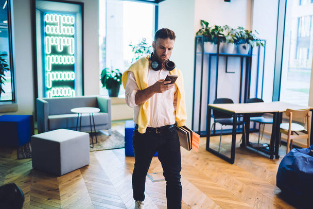 Caucasian male coworker with noise cancellation headphones on neck searching media application on modern cellphone device while checking payment transaction via banking app, technology concept - 写真・画像