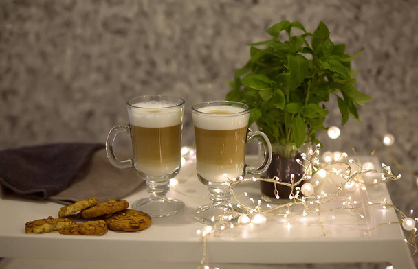 Café con leche de capuchino de chocolate caliente con espuma en una taza de vidrio con galletas. Bandeja blanca con bebidas calientes, guirnalda, bokeh. - Foto, Imagen