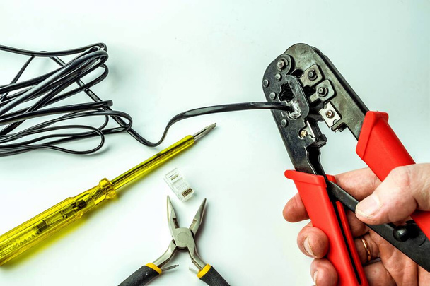 Hand of a craftsman with a crimping tool connecting an RJ45 connector with a black cable on a white background with a screwdriver and pliers. Networks and telecommunications - Valokuva, kuva