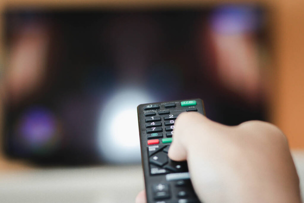 Nahaufnahme der Hand mit dem ferngesteuerten Fernseher und drückt die Taste. Fernsehfernbedienung ändert Kanäle Daumen auf dem blauen TV-Bildschirm. - Foto, Bild