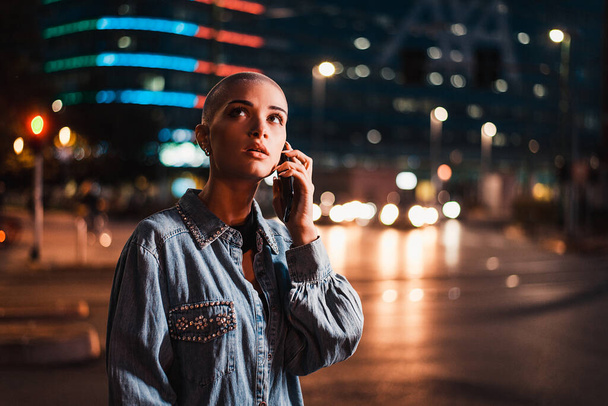 Pretty girl with stylish clothes holding smartphone outdoors in the evening, illuminated city on background - Concepts about technology, communication and lifestyle - Photo, image