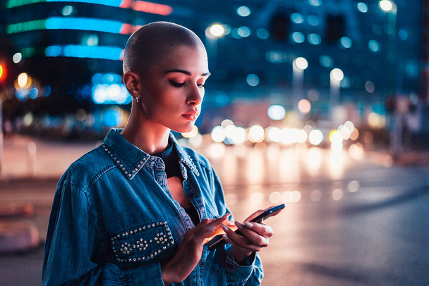 Pretty girl with stylish clothes holding smartphone outdoors in the evening, illuminated city on background - Concepts about technology, communication and lifestyle - Фото, изображение