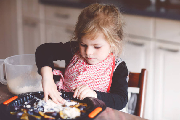 Χαριτωμένο κοριτσάκι που φτιάχνει πίτα δαμάσκηνο στο σπίτι. Χαρούμενο χαμογελαστό παιδί που βοηθάει και ετοιμάζει δαμάσκηνα για κέικ στην οικιακή κουζίνα. Υγιεινό σπιτικό φαγητό. - Φωτογραφία, εικόνα
