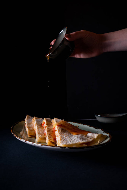 A vertical shot of female hand pouring a sauce into appetizing chicken quesadilla on black background - Foto, Bild