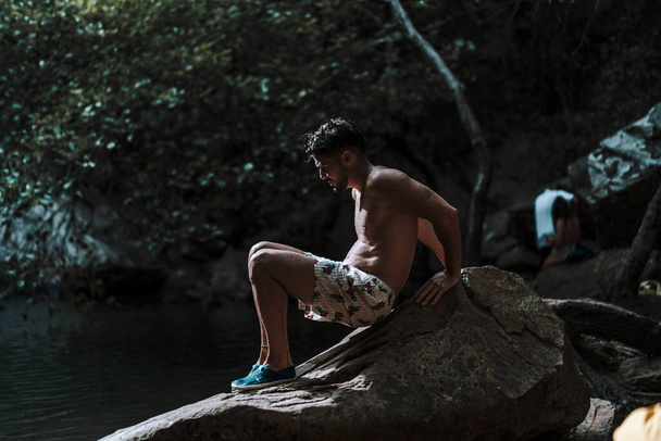 A young Caucasian male sitting on a rock near a river surrounded by beautiful greenery - Fotografie, Obrázek