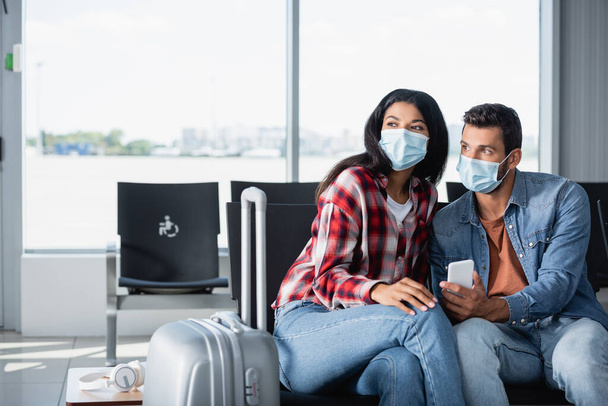 pareja interracial en máscaras médicas sentado cerca de equipaje en la sala de salida  - Foto, imagen