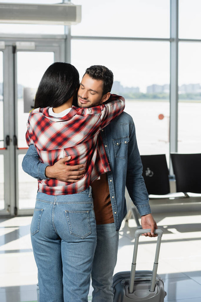 cheerful man smiling while hugging african american woman in airport  - Фото, зображення