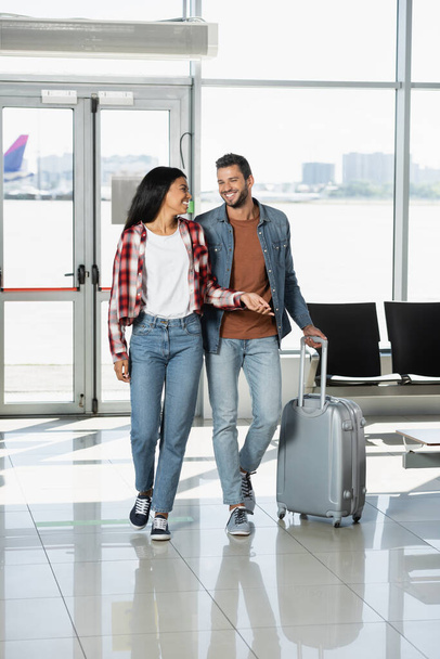 ευτυχισμένο πολυπολιτισμικό ζευγάρι με αποσκευές με τα πόδια στο αεροδρόμιο  - Φωτογραφία, εικόνα