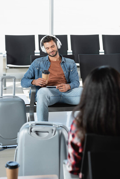 homem barbudo em fones de ouvido segurando copo de papel perto de bagagem e mulher em primeiro plano borrado  - Foto, Imagem