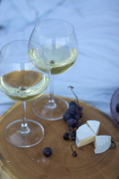 puinen jalusta, jossa on kaksi lasillista samppanjaa, viinirypäleitä ja camembert-juustoa valkoisella pellolla. piknik - Valokuva, kuva