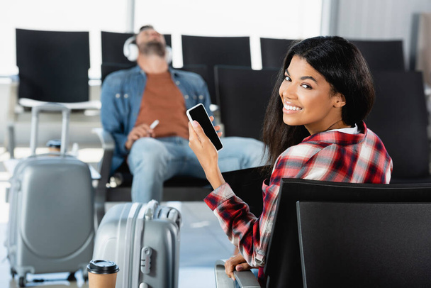 feliz Africano americano mulher segurando smartphone com tela em branco perto de bagagem e homem no fundo borrado  - Foto, Imagem