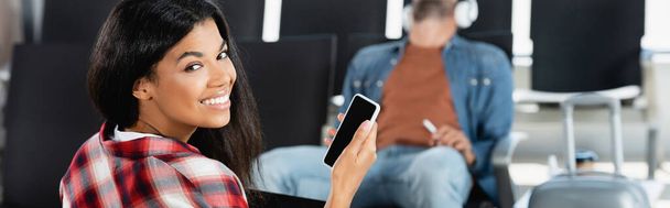 Mutlu Afro-Amerikalı kadın elinde boş ekranlı akıllı telefon, arka planda bulanık bir adam, afiş. - Fotoğraf, Görsel