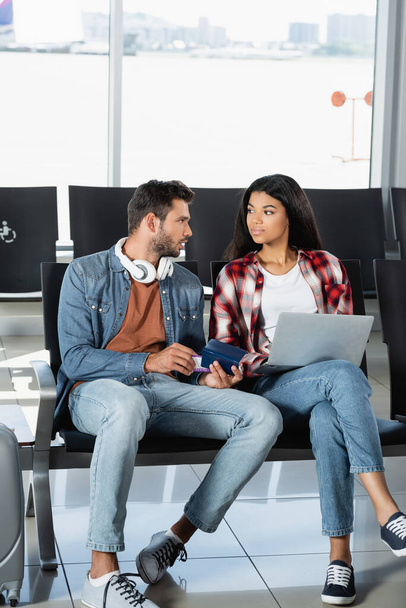 bearded man in headphones holding passport and looking at african american woman with laptop in airport  - 写真・画像