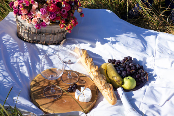 suporte de madeira com dois copos de champanhe, uvas, baguete e queijo camembert em cobertor branco no campo. piquenique - Foto, Imagem
