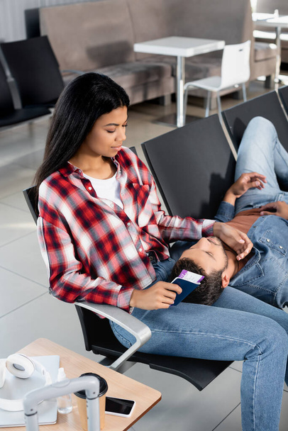 bearded man lying on african american woman with passport in departure lounge of airport - Photo, Image