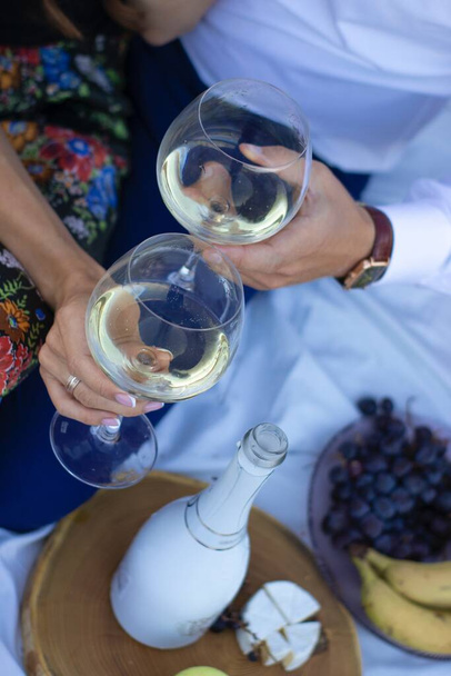 dvě ruce drží sklenice na šampaňské u dřevěného stojanu s lahví, hrozny a sýr camembert na bílé přikrývce. piknik - Fotografie, Obrázek