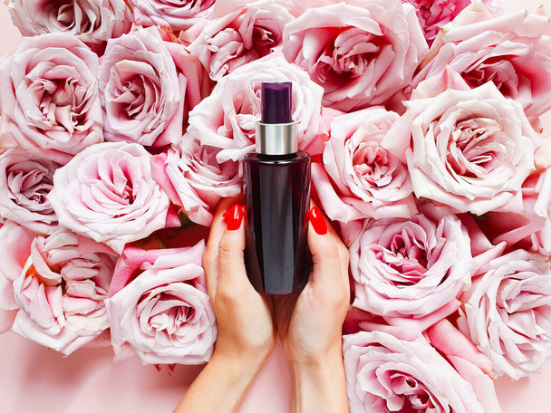 Mockup of dark brown spray bottle, caucasian female hands with red nails and roses on pink table. Top view, copy space - Photo, Image