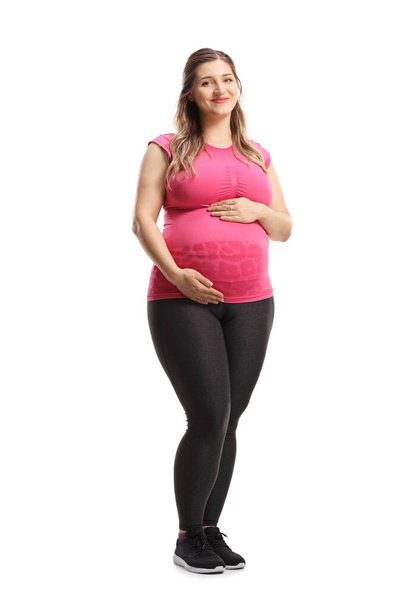 Full length portrait of a pregnant woman in pink top and leggings smiling and holding her belly isolated on white background - 写真・画像
