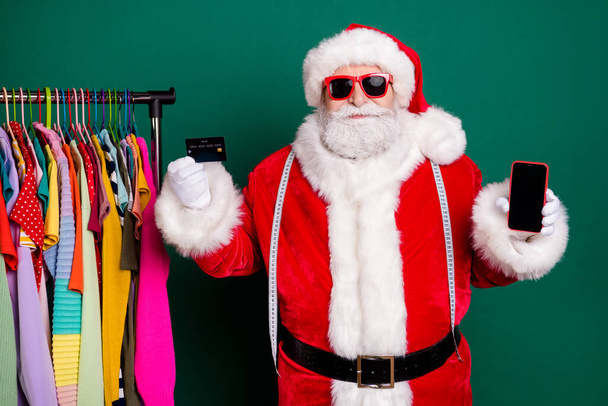 Photo of santa claus showroom boutique shopper buyer enjoy x-mas tradition fairy discount hold smartphone pay credit card wear red cap headwear isolated over green color background - Foto, Imagen