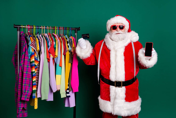 Grey white hair santa claus x-mas enjoy christmas eve boutique shopper buyer discount hold smartphone pay credit card wardrobe closet wear red costume cap isolated green color background - 写真・画像