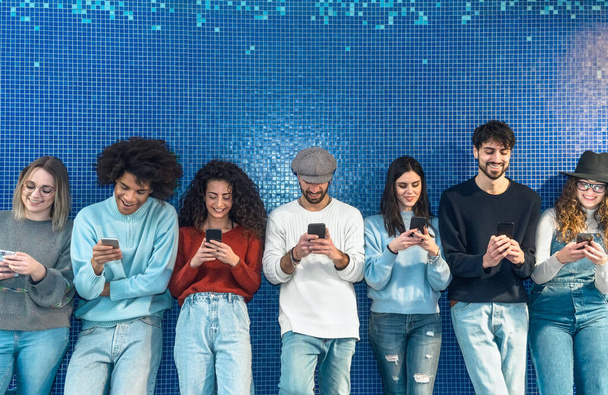 Young people using mobile smartphone while standing against blue background - Youth millennial addicted to new technology social media app concept - Photo, image