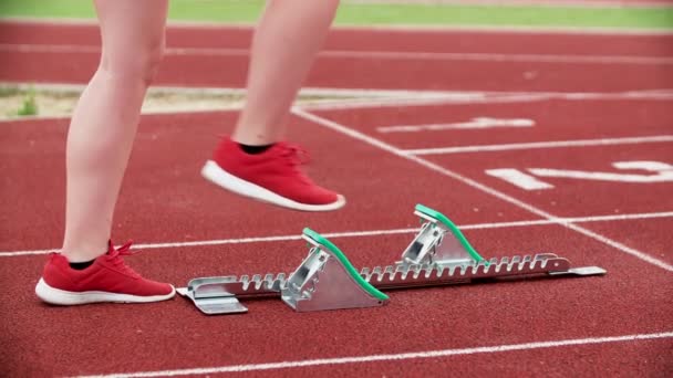 Una joven estudiante en zapatillas rojas va en una posición de partida y luego comenzará a correr en una pista de atletismo. - Metraje, vídeo