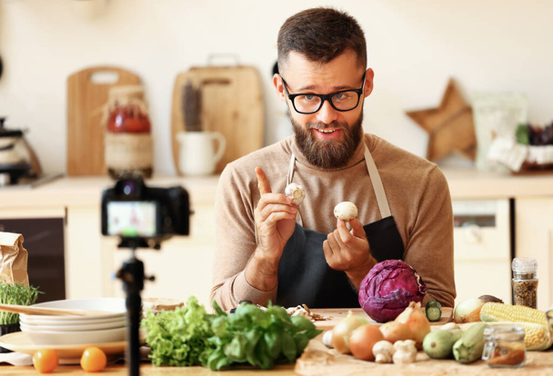 Heureux homme barbu dans des lunettes souriant et démontrant des légumes mûrs tout en tournant une vidéo pour la cuisson vlog dans la cuisine à hom - Photo, image