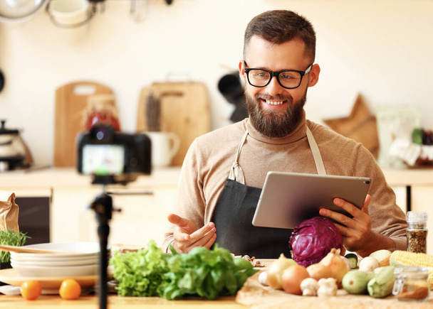 Cheerful young bearded male blogger with tablet in hand sitting at table with fresh organic vegetables and recording video recipe for social networ - Zdjęcie, obraz