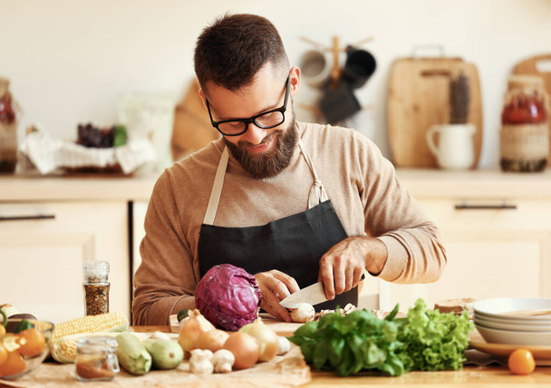 木製のまな板の上にエプロンスライスシャンパンで陽気若いひげそりの男性家庭用キッチンで新鮮な食材と健康的な料理を準備しながら - 写真・画像