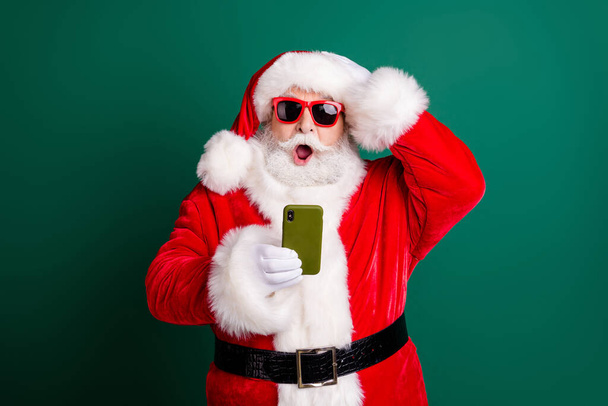 Photo of pensioner stylish grandfather grey hair beard hold telephone amazed open mouth hand head wear red santa x-mas costume coat gloves sunglass cap isolated green color background - Fotó, kép