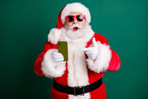 Photo of pensioner stylish grandfather grey hair beard hold directing finger telephone assure quality wear red santa x-mas costume coat gloves sunglass cap isolated green color background - 写真・画像
