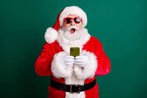 Photo of pensioner stylish grandpa grey hair beard hold telephone shocked open mouth crazy novelty wear red santa x-mas costume coat gloves sunglass headwear isolated green color background - Fotografie, Obrázek