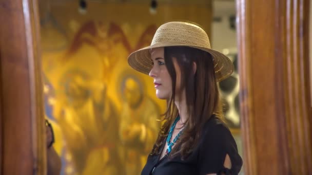 博物館の若い女性は素敵なわらの帽子をしようとしており、彼女は非常にエレガントに見えます。鏡の中を見てる. - 映像、動画