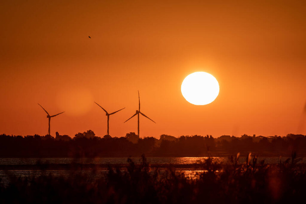 Generadores de viento silueta granja en la costa del mar en el amanecer rojo naranja cielo vibrante y gran sol naciente con brillo en el agua. Turbinas energéticas industria sostenible - Foto, Imagen