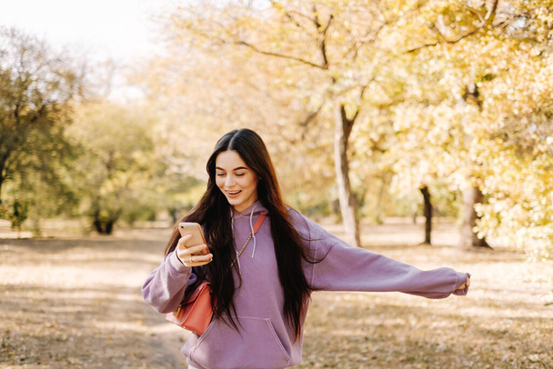 Beautiful young woman having fun, using smartphone and enjoying autumn weather in the park. Woman walking in the autumn park. High quality photo - Photo, Image