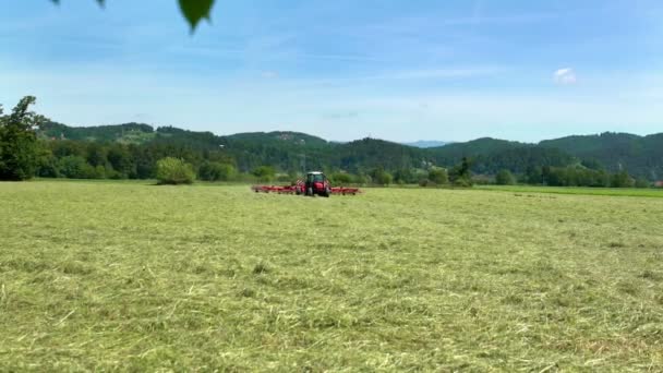 De natuur is prachtig op deze zomerdag. Een blauwe tracto bereidt hooi voor en trekt er grote landbouwmachines achter aan.. - Video