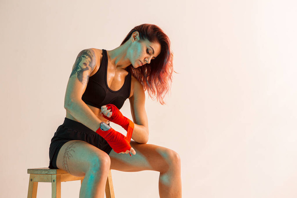 A young boxer girl with red bandages on her hands poses on a chair in a photo Studio. - Фото, изображение