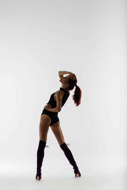 Young beautiful flexible female in a black jumpsuit and high heels is posing in a dance studio. - Foto, afbeelding