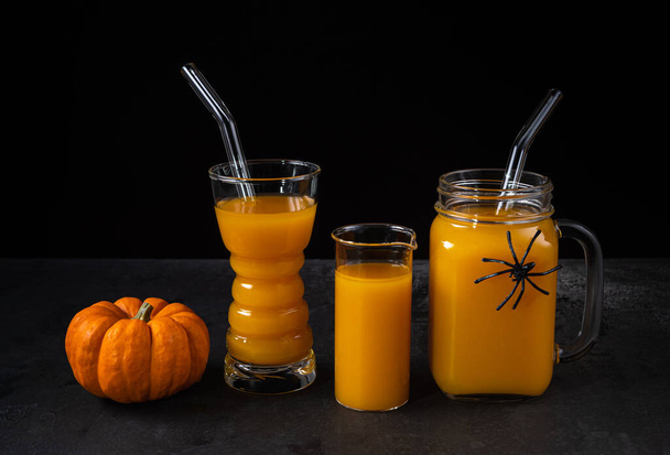 Non-alcoholic pumpkin mocktail in glass, measuring glass and square jar with glass straws and whole orange pumpkin on black textured surface. Halloween spider decor on jar. Healthy eating concept. - Fotografie, Obrázek