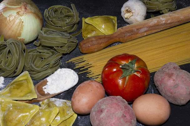 Pâtes alimentaires fond avec un espace de copie de raviolis, spaghettis, oeuf, pomme de terre, tomate et un pétrin de bois sur fond noir farineux. - Photo, image
