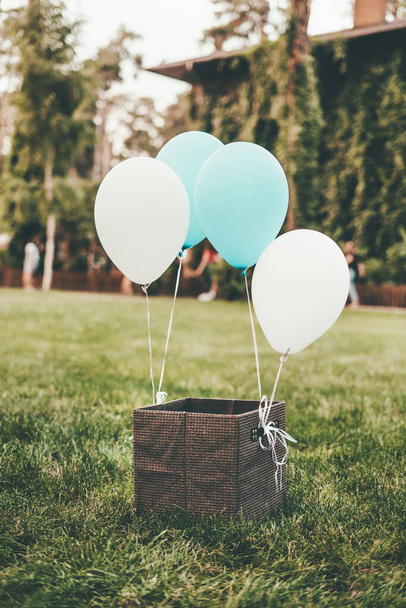 Eine braune Schachtel steht auf grünem Gras mit gebundenen weißen und blauen Luftballons.  - Foto, Bild