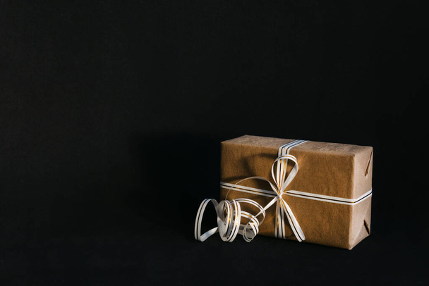 Gift box wrapped in recycled craft paper and tied with a white and gold ribbon bow on a black background. Surprise for the holiday. - Фото, зображення