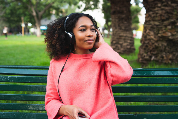 Retrato da jovem afro-americana ouvindo música com fones de ouvido e celular no parque. Ao ar livre. - Foto, Imagem