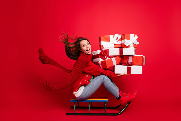Full length body size profile side view of attractive glad cheerful girl elf Santa assistant riding sledge delivering giftboxes bargain isolated bright vivid shine vibrant red color background - Photo, image