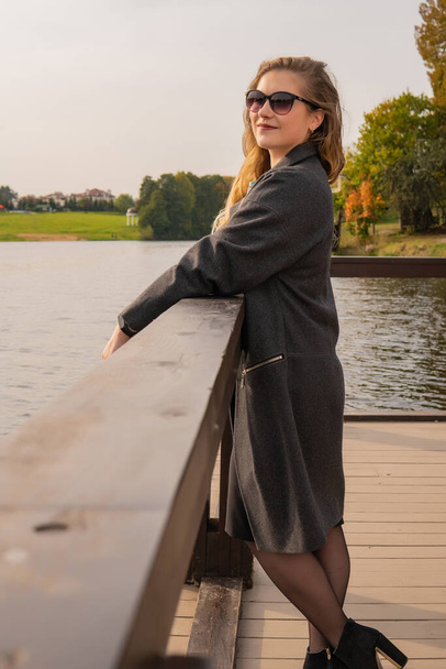 Cheerful young woman standing near the water with a charismatic appearance with a Caucasian appearance in a coat in the fall on the pier in the afternoon - Фото, изображение