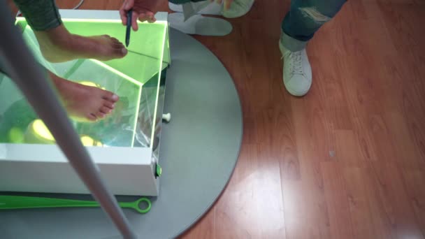 Paciente femenina descalza que permanece en superficie de vidrio transparente con luz de neón verde mientras el médico ortopedista examina las condiciones de los pies y los huesos. Médico explicando el problema de pie plano y - Metraje, vídeo